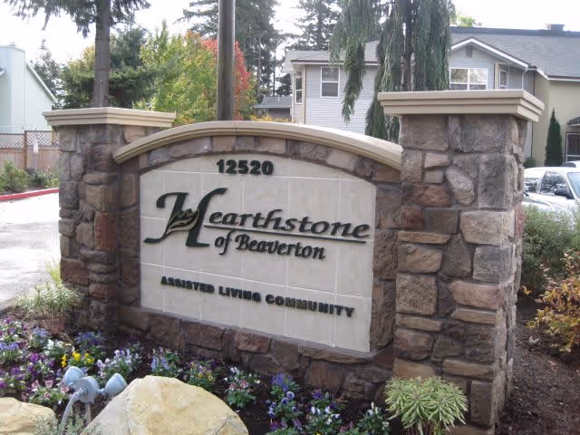 Stone sign at the entrance of Hearthstone of Beaverton assisted living community with the address 12520, surrounded by flowers and plants, with a residential building and parked cars in the background.