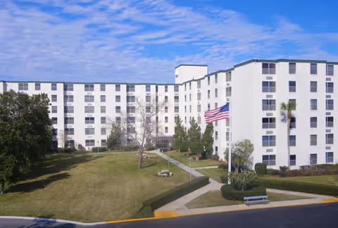 Exterior view of a large white multi-story senior living facility building with multiple windows. There is a well-maintained lawn with trees, a picnic table, a bench, and an American flag on a flagpole in front of the building under a partly cloudy blue sky.