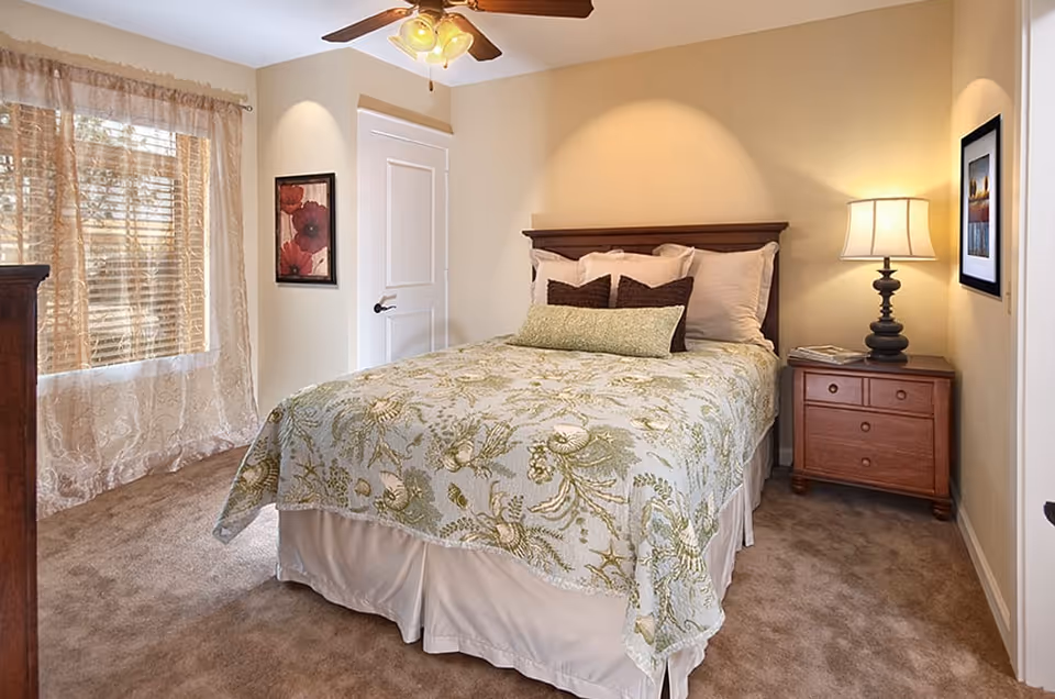 A cozy bedroom with a double bed covered in a green and beige floral bedspread. The bed has multiple pillows and is positioned against a beige wall. To the right of the bed is a wooden nightstand with a lamp and some magazines. There are two framed pictures on the walls, one above the nightstand and one near the door. A window with sheer curtains allows natural light to enter the room. The floor is carpeted in a light brown color, and a ceiling fan with lights is mounted above.