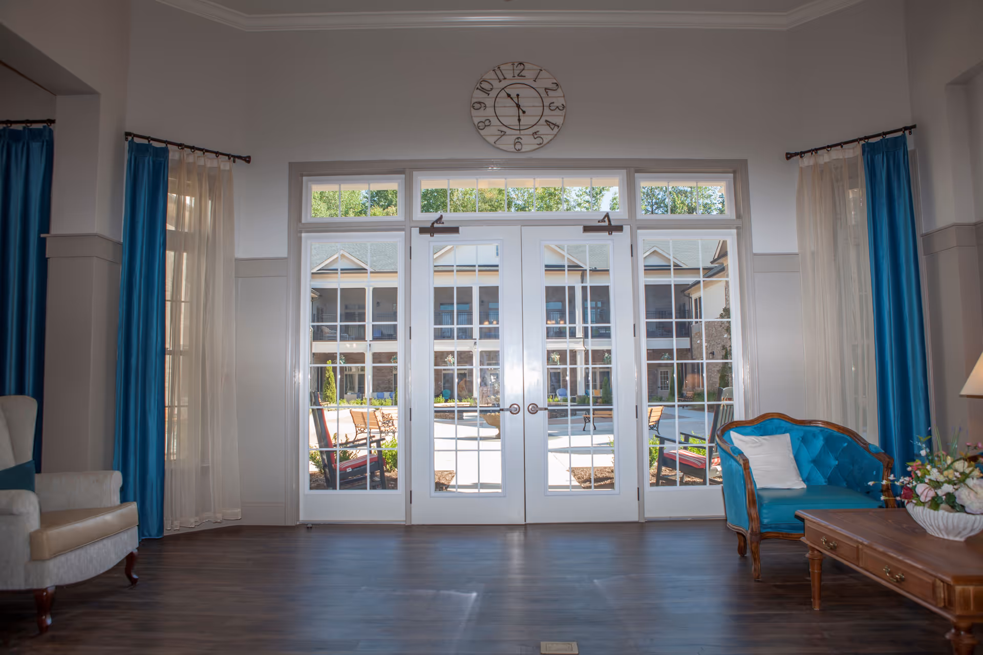Interior view of a senior living facility lounge area with large glass double doors leading to an outdoor courtyard. The room features blue curtains, a beige armchair on the left, a blue upholstered chair with a white pillow on the right, and a wooden coffee table with a floral arrangement. A large clock is mounted above the doors.