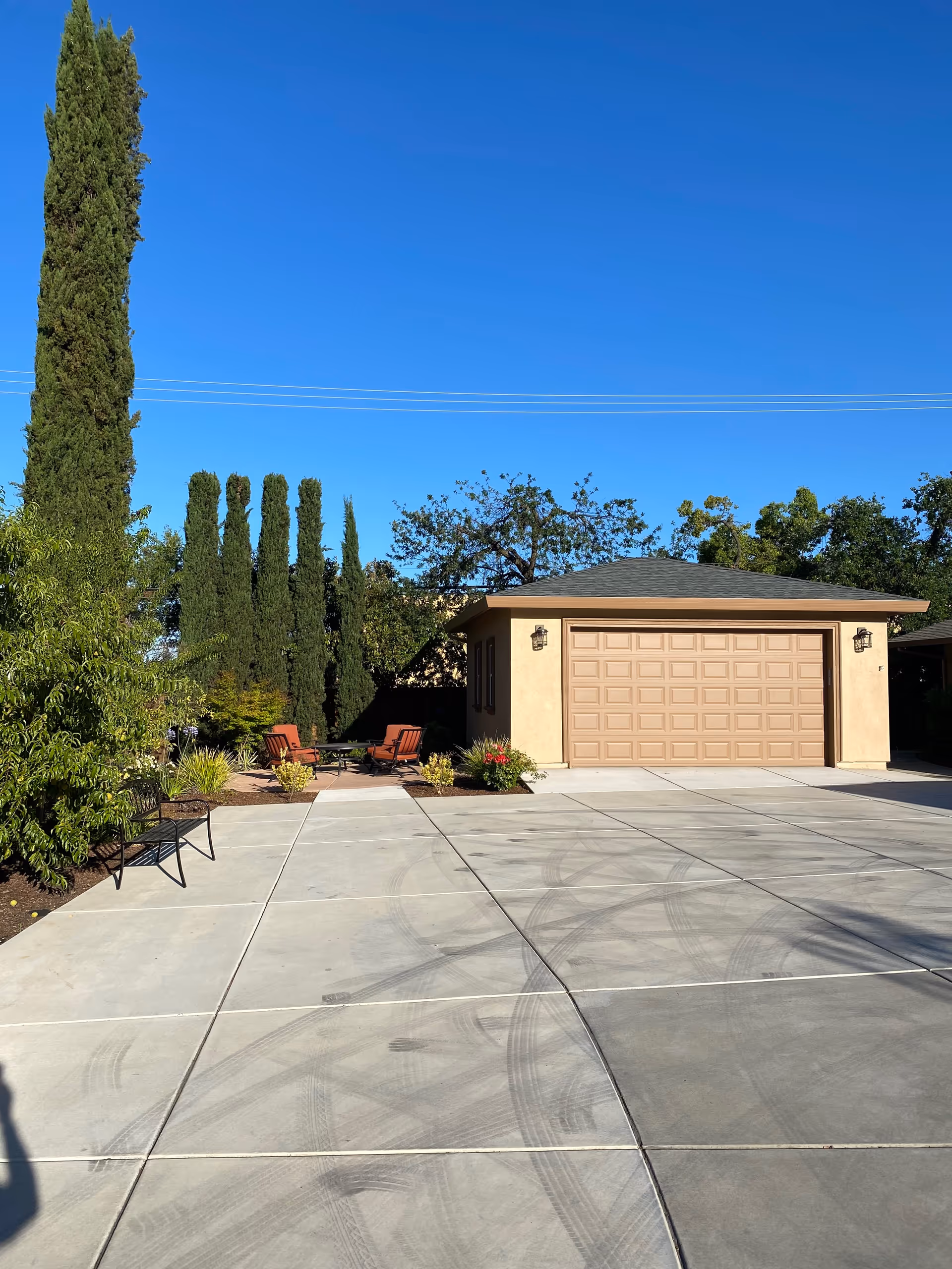 Outdoor view of a paved driveway leading to a beige garage with a closed door. To the left, there are tall, narrow trees and some shrubs, along with a bench and a seating area with chairs and a small table. The sky is clear and blue.