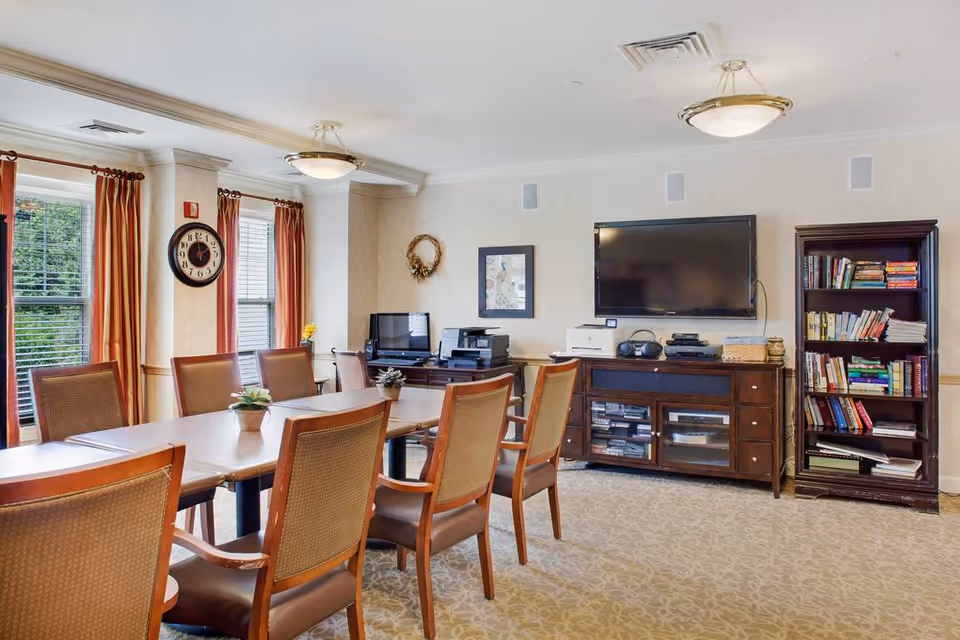 Bright communal dining/activity room with a long table surrounded by chairs, a wall-mounted TV, bookshelf, and windows with curtains.