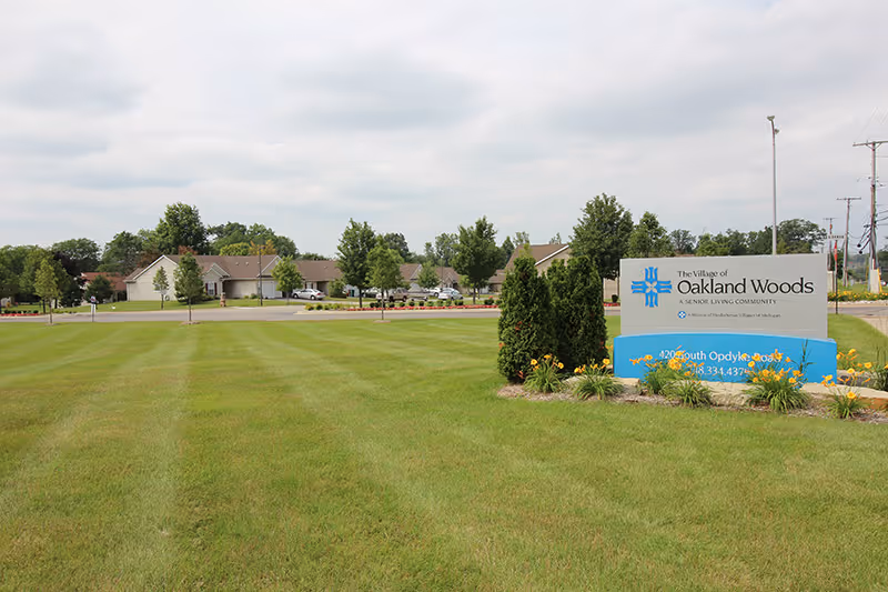 A large green lawn with a sign for The Village of Oakland Woods, a senior living community. The sign is surrounded by small bushes and flowers. In the background, there are single-story buildings and trees under a cloudy sky.