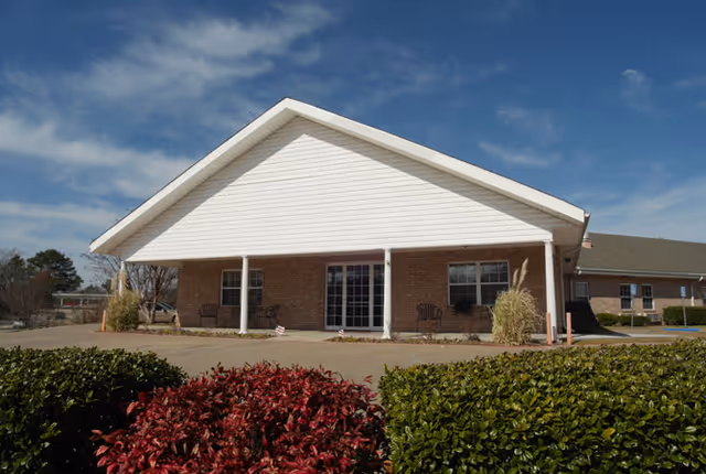 Front exterior view of a single-story brick building with a white gabled roof and a covered porch area. There are two chairs on the porch, and neatly trimmed bushes and red-leaved plants in the foreground under a blue sky with some clouds.