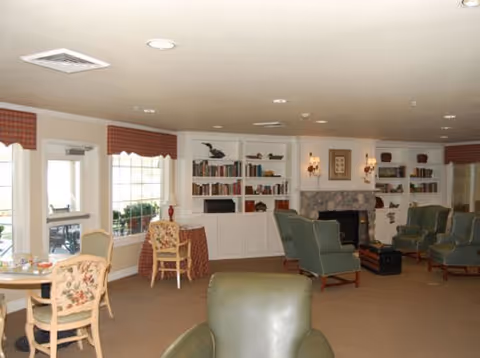 A cozy living room area with green upholstered chairs arranged around a stone fireplace. Built-in white bookshelves flank the fireplace, filled with books and decorative items. There are windows with red valances letting in natural light, and a small round table with floral upholstered chairs near the windows.