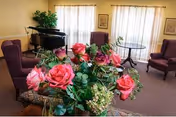 A cozy living room area with a bouquet of pink and red roses in the foreground. The room features purple armchairs, a black grand piano, framed artwork on the walls, and large windows with sheer white curtains allowing natural light to fill the space.
