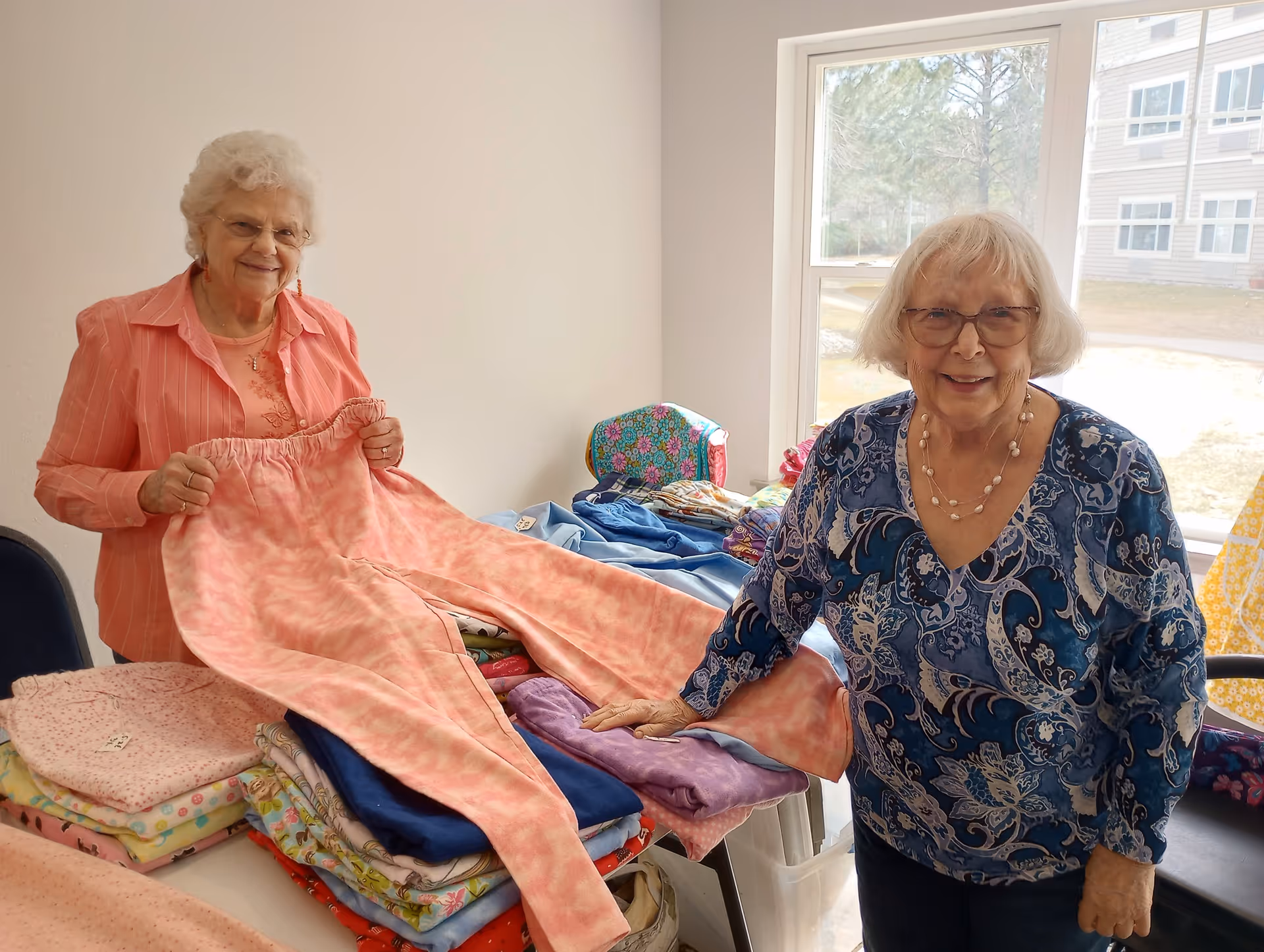 Two elderly women standing indoors near a table covered with folded colorful fabrics and clothing. One woman is holding up a pair of pink pants, and both are smiling. A window in the background shows an outdoor view with trees and a building.