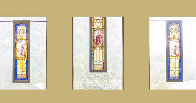 Three vertical stained glass windows with religious imagery, set against a beige wall with a view of trees outside.