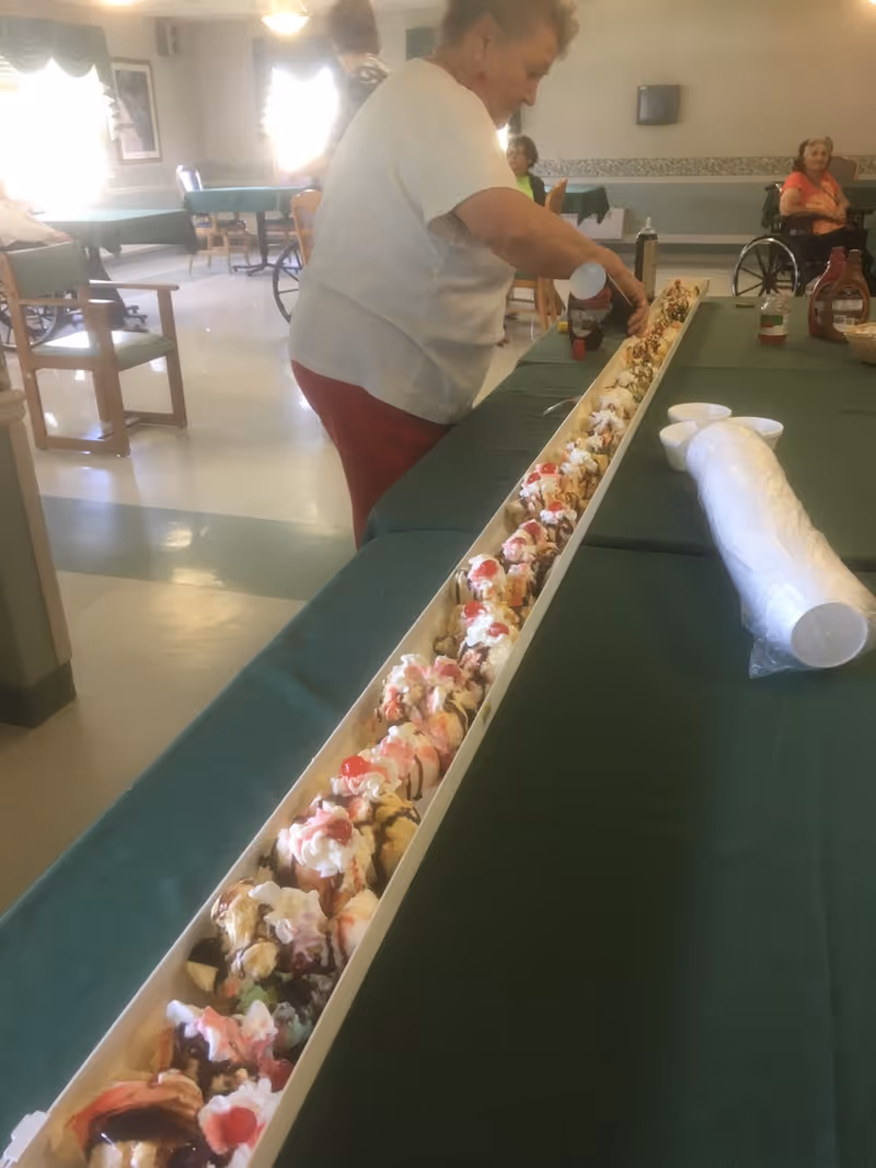 An elderly woman is adding toppings to a very long tray of ice cream sundaes lined up on a green tablecloth in a communal dining area. Other elderly individuals are seated in the background, some in wheelchairs. The room has tables and chairs arranged for dining.