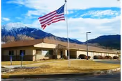 Single-story beige building with American flag, parking area, and mountain backdrop.