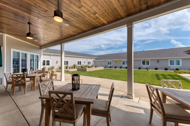 Covered outdoor patio area with wooden ceiling and ceiling lights, furnished with wooden tables and cushioned chairs. Beyond the patio is a grassy courtyard surrounded by a single-story building with multiple windows under a partly cloudy sky.