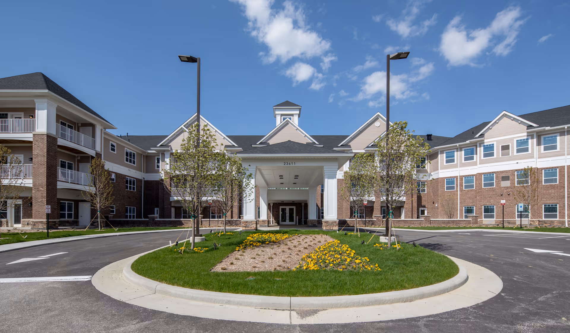 Front entrance of a multi-story senior living building with a circular driveway, landscaped center island and lamp posts under a blue sky.