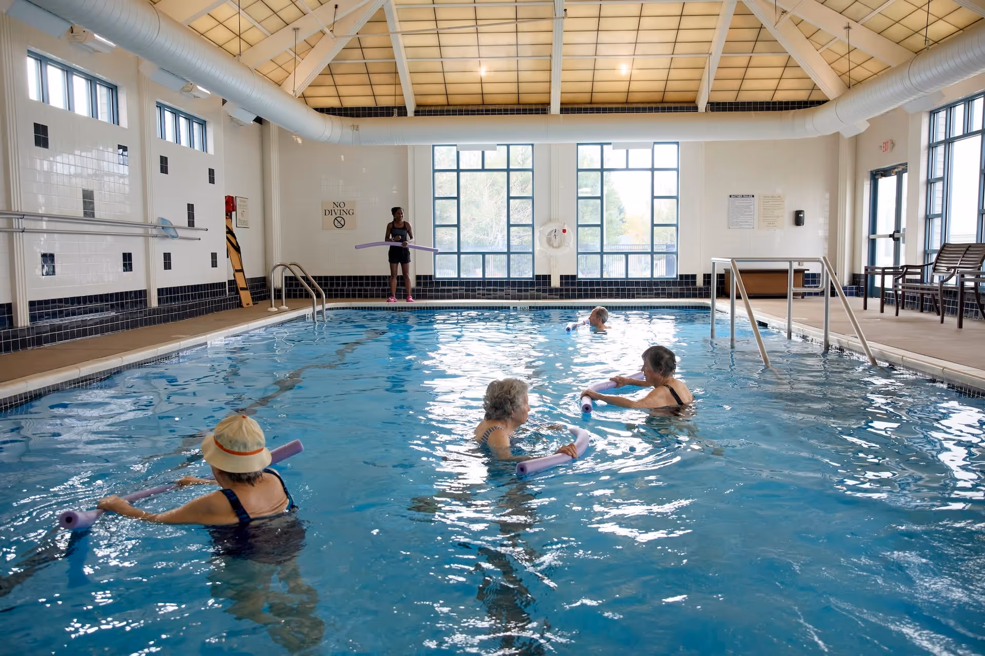 Indoor swimming pool with four elderly women in the water holding purple pool noodles and a young instructor standing at the poolside holding a pool noodle. The pool area has large windows, white tiled walls with black accents, and a high ceiling with exposed ducts.