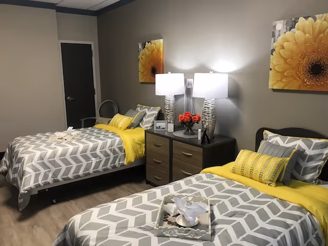 A cozy bedroom with two twin beds featuring gray and white chevron patterned bedding and yellow accents. Between the beds is a wooden nightstand with two modern silver lamps, a small vase of red flowers, and a framed photo. The walls are painted gray and decorated with yellow flower artwork. The floor is light wood, and there is a closed door in the background.