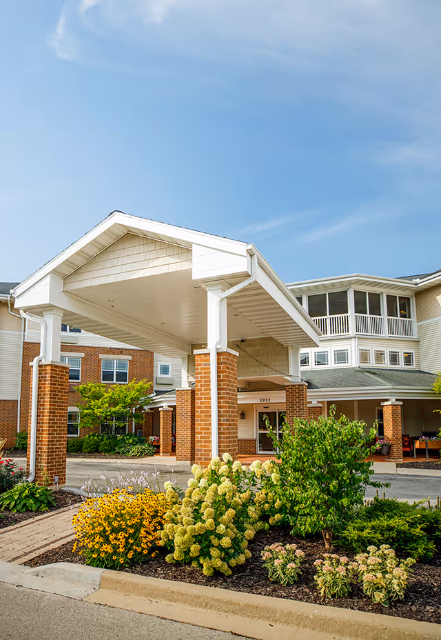 Entrance of Hickory Park Senior Living facility with a covered drop-off area supported by brick columns, surrounded by well-maintained landscaping with various flowering plants and shrubs under a clear blue sky.