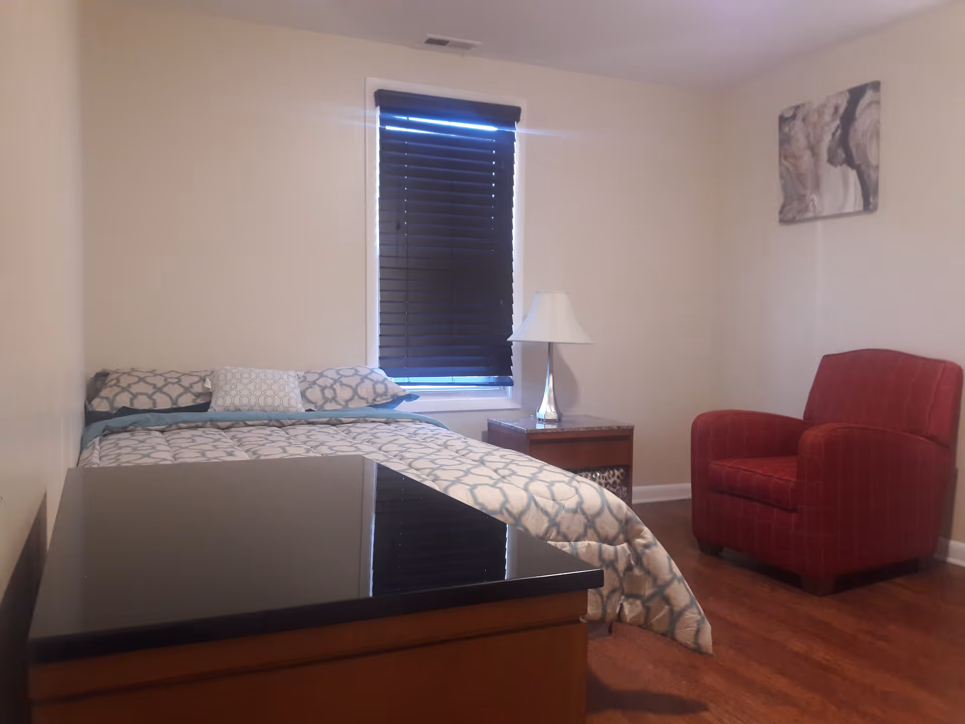 Small bedroom with a bed, nightstand and lamp, a red armchair, and a window with closed blinds.