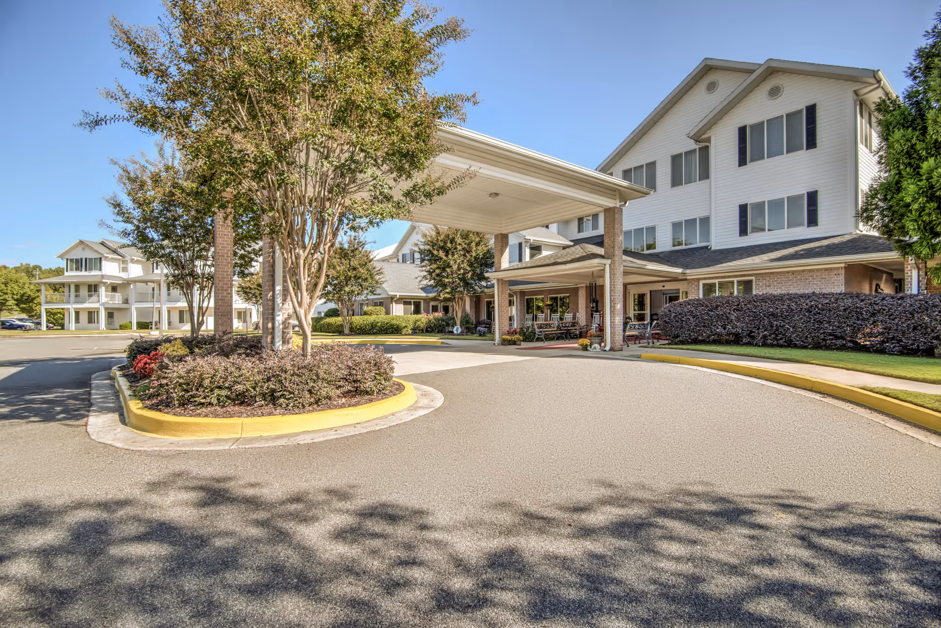 Exterior front entrance of a multi-story senior living building with a covered porte-cochere, circular driveway, and landscaped beds.