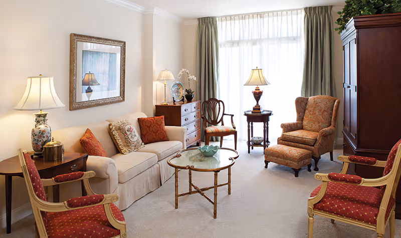 A cozy living room with a beige sofa adorned with decorative pillows, two red upholstered armchairs with wooden frames, a patterned armchair with matching ottoman, a glass-top coffee table, a wooden chest of drawers with a lamp and decorative items, and a large window with sheer curtains allowing natural light to fill the room.