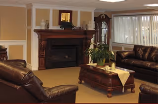 A cozy living room with two dark brown leather sofas facing a wooden fireplace with a decorative mantel. A wooden coffee table with a yellow runner and a potted plant sits in the center. Large windows with vertical blinds allow natural light into the room.