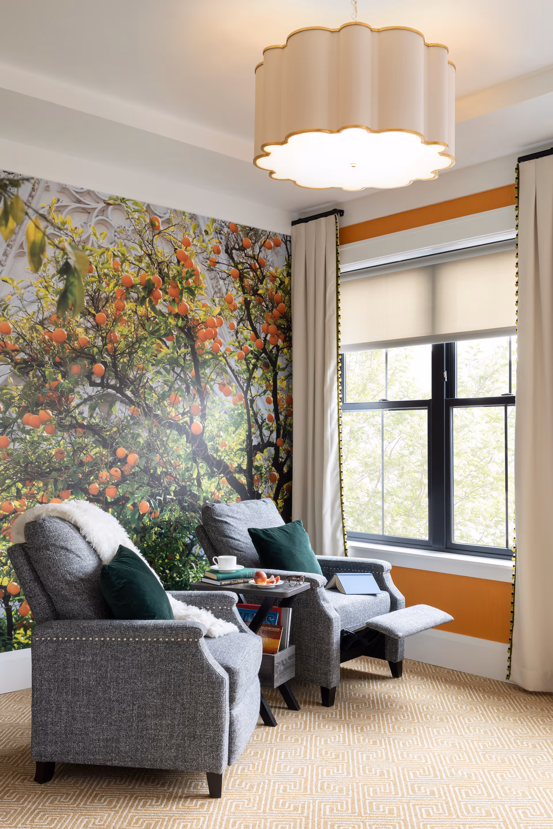 Two gray recliners with green pillows and a small side table by a window in a cozy lounge with an orange-tree mural and a scalloped ceiling light.