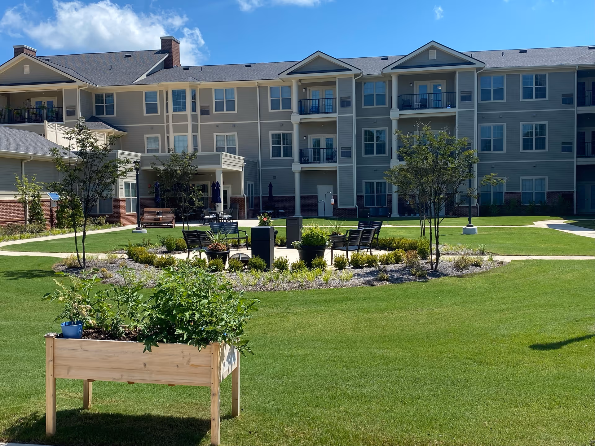 Outdoor courtyard area of a senior living facility with green grass, small trees, a raised wooden planter box with plants, and a circular seating area with chairs and potted plants. The background shows a multi-story building with balconies and windows under a clear blue sky.