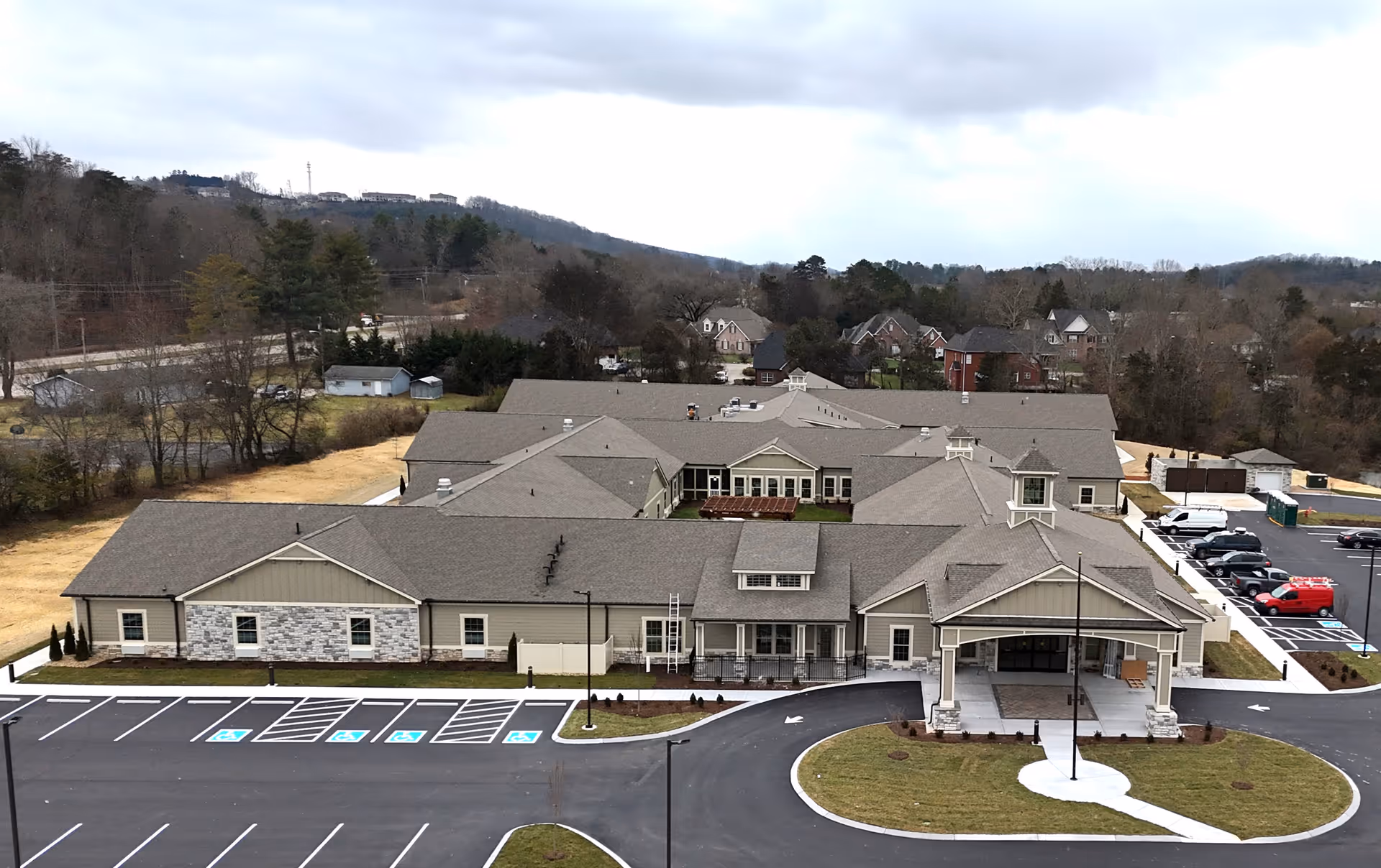 Aerial view of a large single-story senior living facility building with a covered entrance, multiple roof sections, and a spacious parking lot with several handicap parking spaces. The building is surrounded by some trees and residential houses in the background under a cloudy sky.