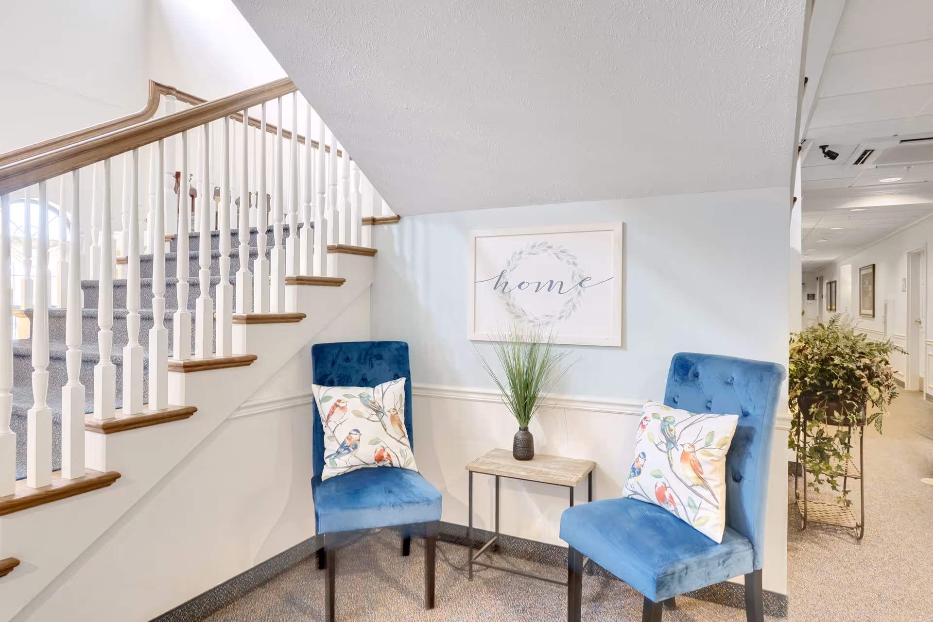 A cozy seating area with two blue velvet chairs featuring bird-patterned pillows, a small wooden table with a vase of green plants, and a framed wall art with the word 'home' above the table. The area is next to a carpeted staircase with white railings and wooden handrails, and a hallway with plants and framed pictures is visible in the background.