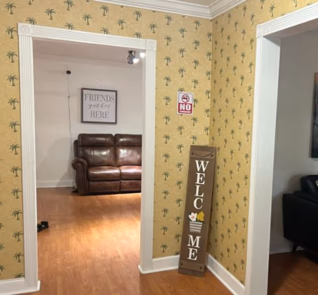 Interior view of a senior living facility showing a hallway with yellow wallpaper featuring small palm tree patterns. A vertical wooden sign leaning against the wall reads 'WELCOME' with decorative flowers. Through an open doorway, a brown leather couch is visible in a room with wooden flooring and a framed sign on the wall that says 'FRIENDS get close HERE'. Another room with a black chair is partially visible on the right side.
