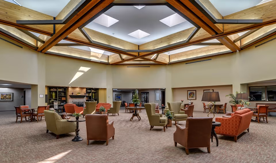 A spacious senior living facility common area with multiple seating arrangements including armchairs and sofas in earth tones. The room features a high ceiling with wooden beams and skylights allowing natural light to fill the space. There are side tables with plants and lamps, and a fireplace with built-in bookshelves in the background.