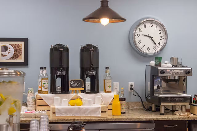 A coffee and beverage station with two large black coffee dispensers, a small chalkboard sign that says 'Add Flavor', several bottles of flavor syrup, a coffee machine, a container with lemon slices, disposable cups, and a wall clock showing the time as 10:50.