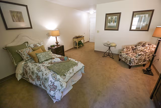 A cozy bedroom with a single bed adorned with patterned bedding and multiple pillows. Next to the bed is a nightstand with a lamp and an alarm clock. Across from the bed is a floral upholstered armchair beside a small round glass-top table with decorative items. The room has beige carpet, white walls, framed artwork, and a floor lamp in the corner. A small console table with plants is visible near the doorway.