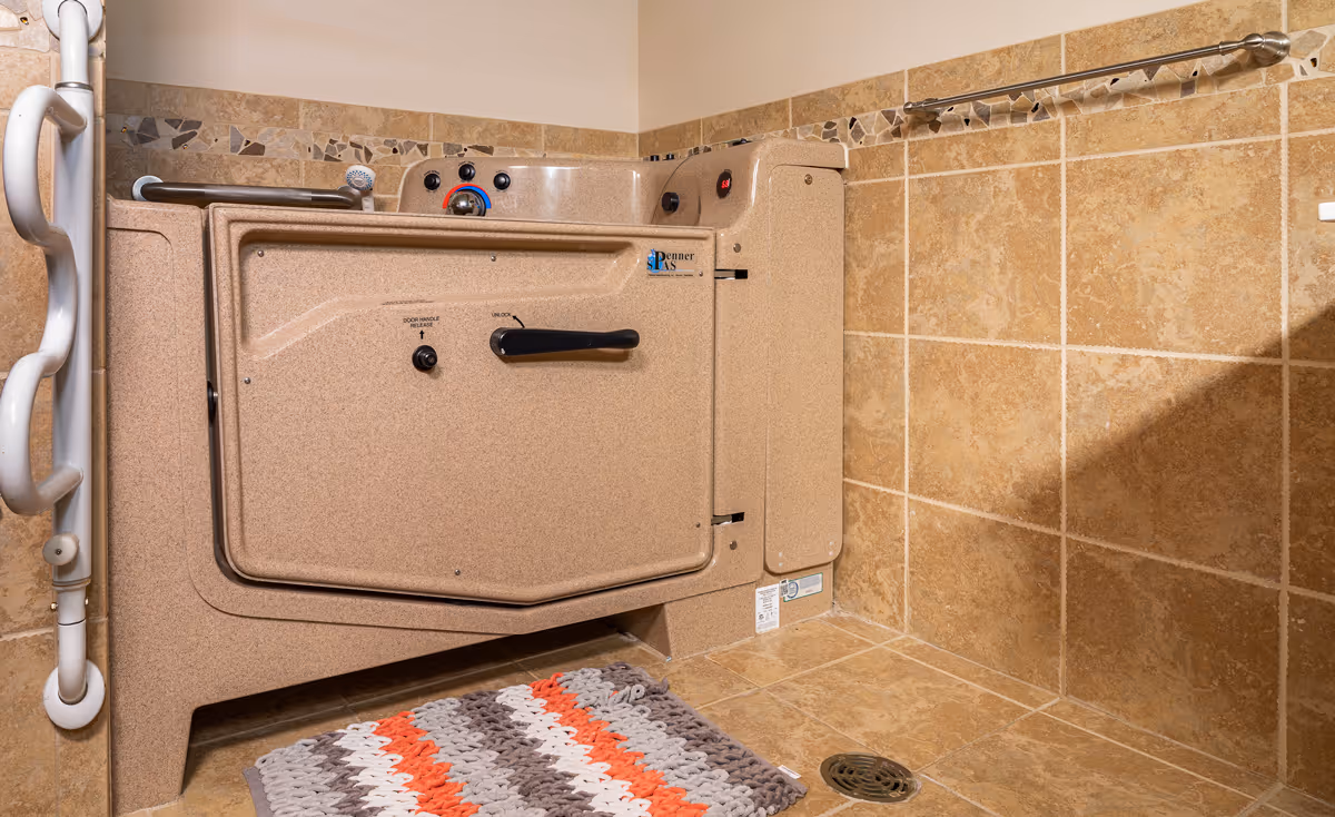 A beige walk-in bathtub with safety handles and control knobs, surrounded by tan tiled walls and floor. A striped bath mat is placed in front of the tub, and a metal towel bar is mounted on the wall.