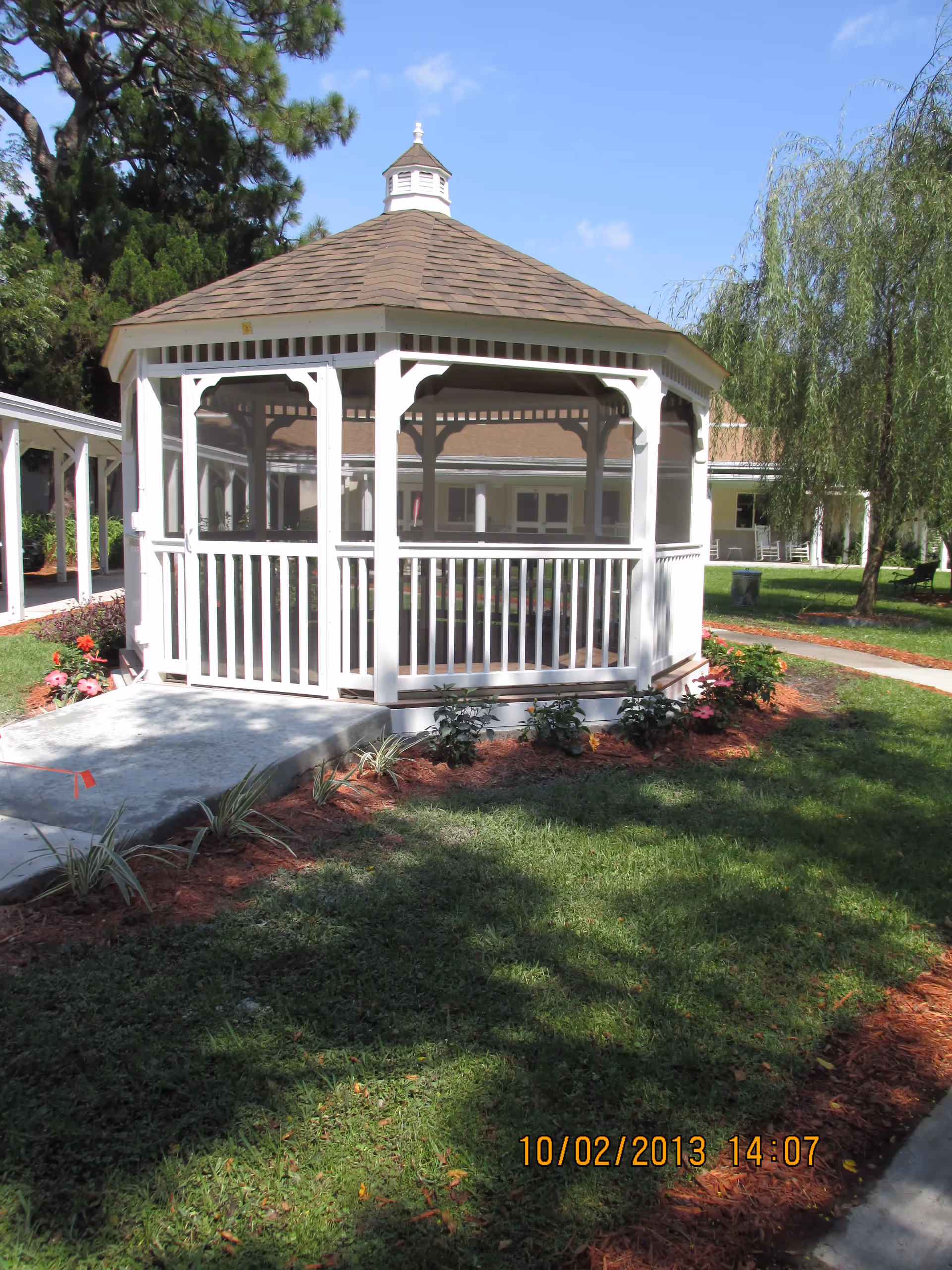 White octagonal gazebo on a grassy courtyard with landscaping and a building in the background.