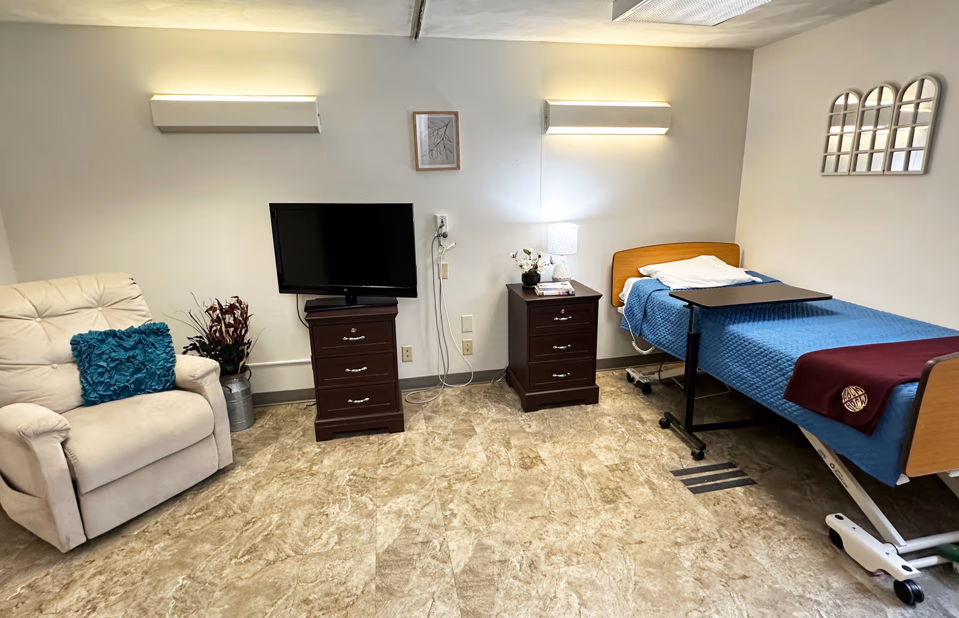 A senior care facility room with a beige recliner chair with a teal cushion, a flat-screen TV on a dark wooden dresser, a matching nightstand with a lamp and decorative flowers, and a hospital-style bed with blue bedding and a maroon blanket. The room has beige walls, two wall-mounted lights, and a small framed picture above the TV.