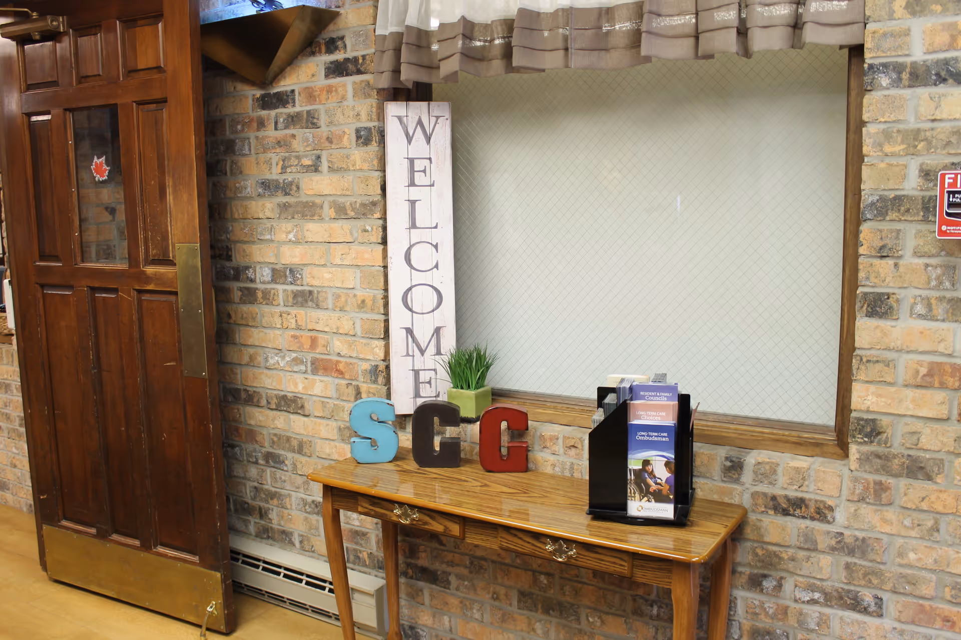A wooden table against a brick wall with a vertical 'WELCOME' sign, decorative letters 'S', 'C', and 'G', a small green plant, and a brochure holder containing pamphlets. To the left is a partially open wooden door with a brass kick plate, and to the right is a window with a beige curtain and a fire alarm sign on the wall.