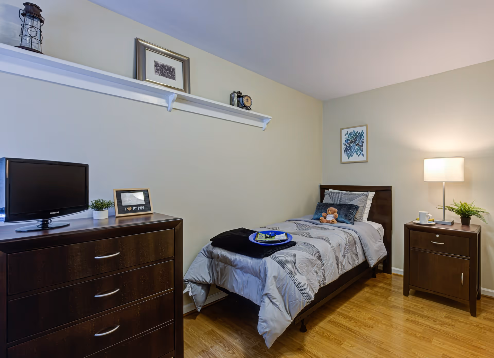 A small, neatly arranged bedroom with a single bed covered in gray and white bedding, a teddy bear on the bed, and a tray with glasses and a book. To the right of the bed is a wooden nightstand with a lamp, a plant, and a cup. To the left is a dark wooden dresser with a small TV, a framed photo, and a small plant. Above the bed and dresser is a white shelf with decorative items and a framed picture on the wall. The floor is wooden.