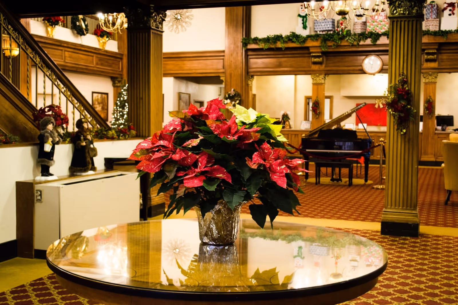 Interior view of a senior residence lobby decorated for the holidays with a round wooden table holding a large poinsettia plant in the foreground. The background features wooden columns, a grand piano, festive garlands, and holiday figurines near a staircase.