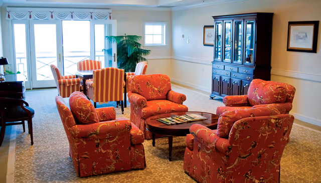 A cozy living room area with four red patterned armchairs arranged around a wooden coffee table with magazines on it. Behind the seating area, there is a dining table with four striped chairs near large glass doors letting in natural light. A dark wooden cabinet with glass doors and framed pictures on the wall are visible in the background.