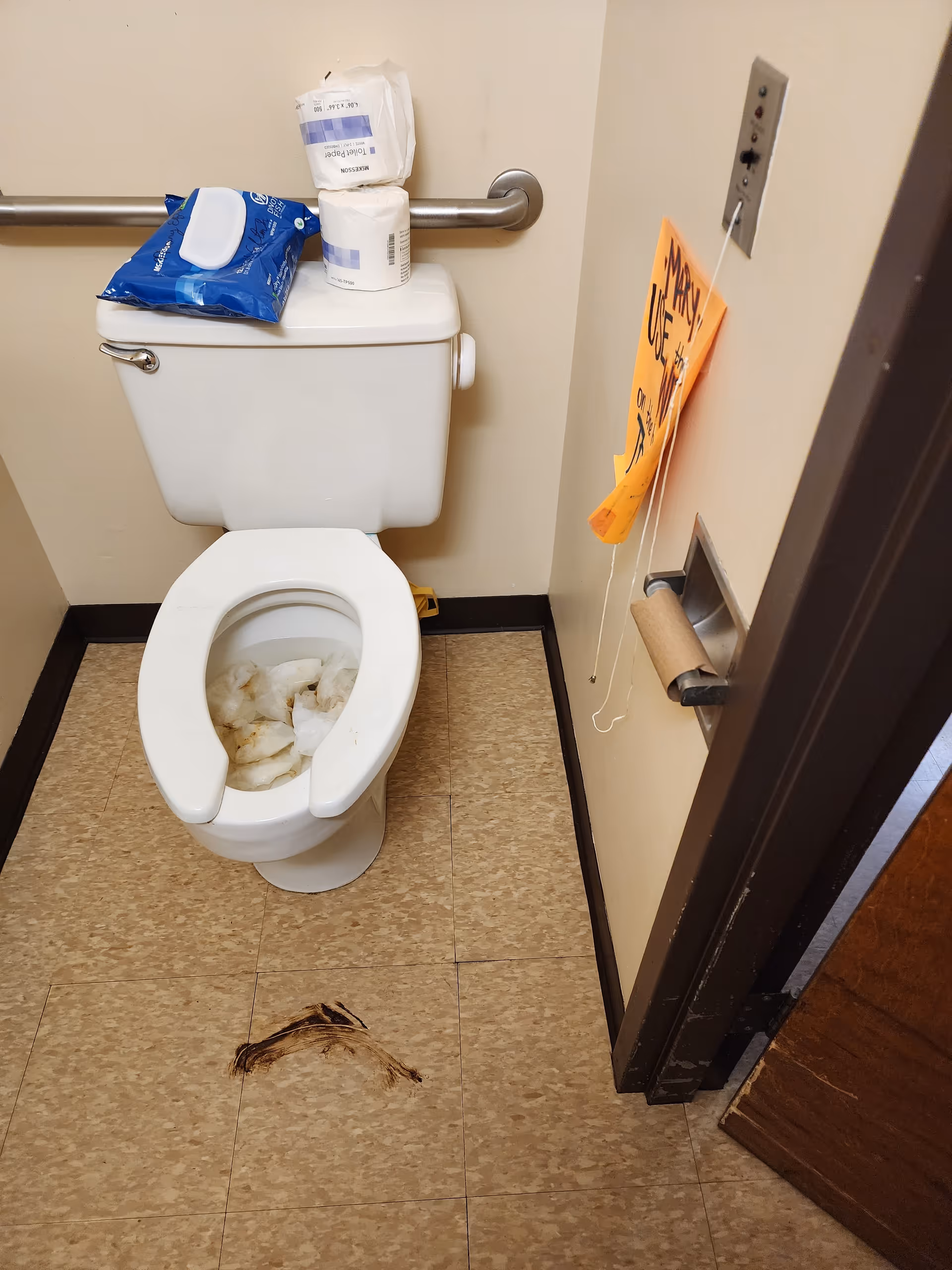 A small bathroom with a white toilet that has used toilet paper inside the bowl. On top of the toilet tank, there are two rolls of toilet paper and a pack of wet wipes. The floor has a dark stain in front of the toilet. There is a metal grab bar on the wall behind the toilet and an empty toilet paper holder on the right wall with an orange sign hanging above it.