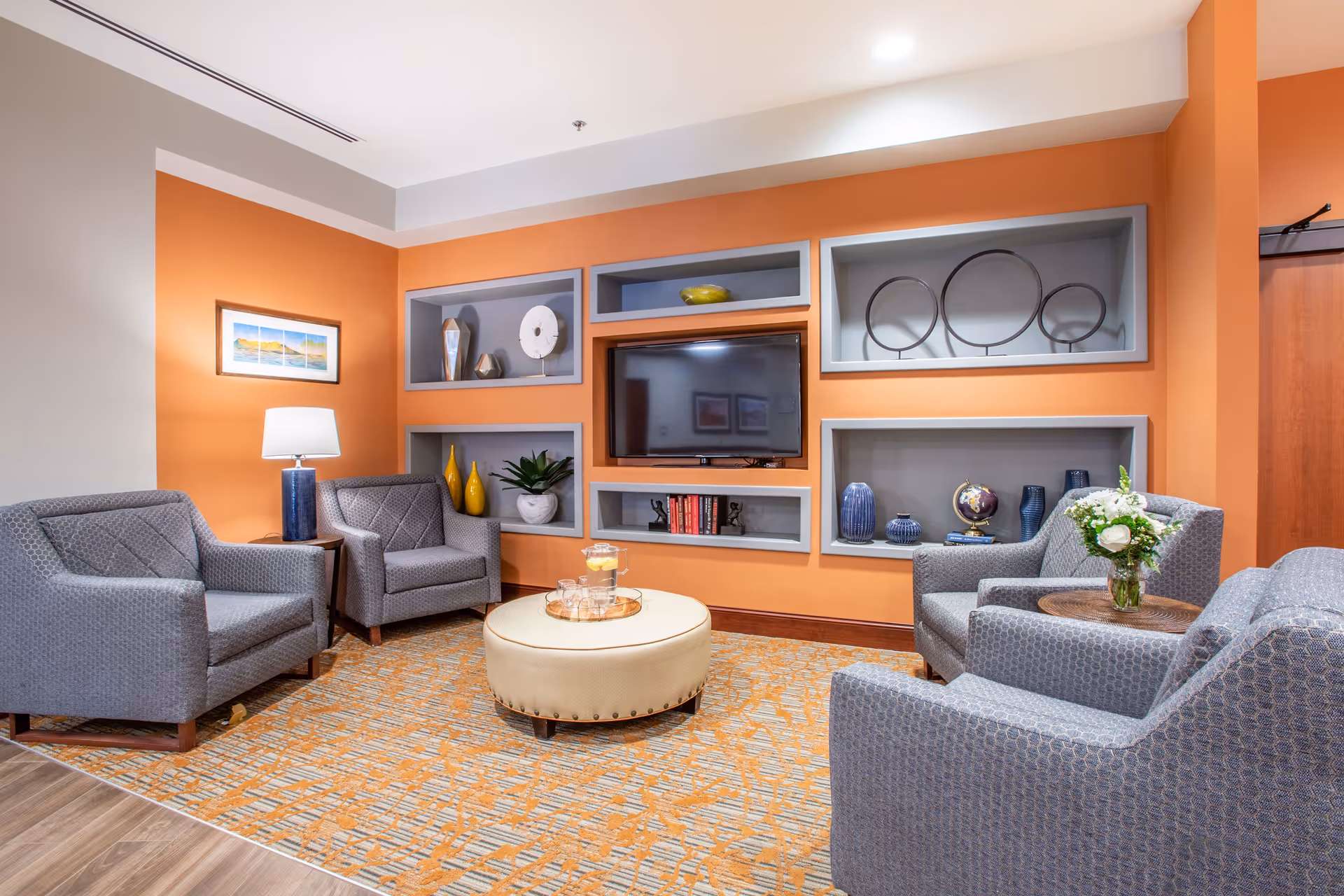 Comfortable seating area with four gray armchairs arranged around a round ottoman in front of a recessed TV and decorative shelves on an orange wall.
