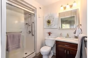 A clean and modern bathroom featuring a glass-enclosed shower with a towel hanging on the handle, a toilet, and a vanity with a sink, mirror, and three-light fixture above. There is a decorative wall art piece above the toilet and a towel hanging on a hook beside the vanity.