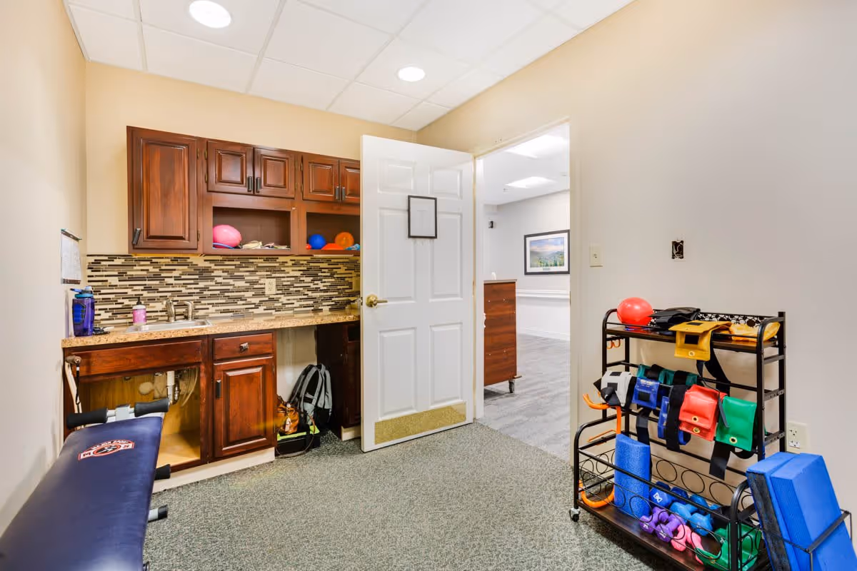 A small room with beige walls and carpeted floor featuring a wooden cabinet with a granite countertop and a mosaic tile backsplash. The cabinet has an open space underneath with plumbing visible. There is a blue exercise bench on the left side and a black metal rack on the right holding various colorful exercise equipment such as weights, resistance bands, and yoga blocks. A white door is open, showing a glimpse of a hallway with gray flooring and a framed picture on the wall.