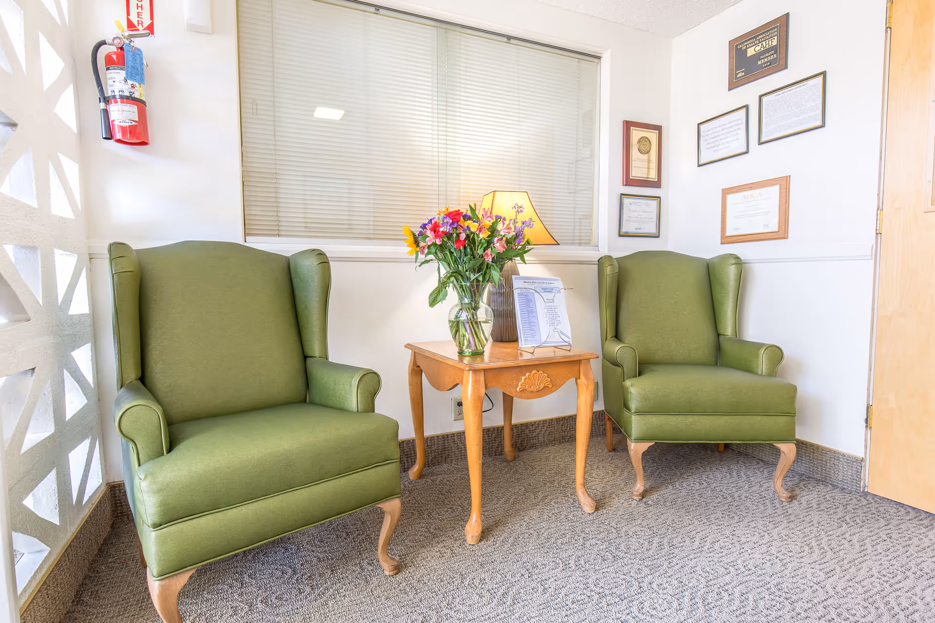 A small seating area with two green upholstered armchairs separated by a wooden side table holding a vase of colorful flowers and a lamp. The wall behind has framed certificates and a window with closed blinds. A fire extinguisher is mounted on the left wall.