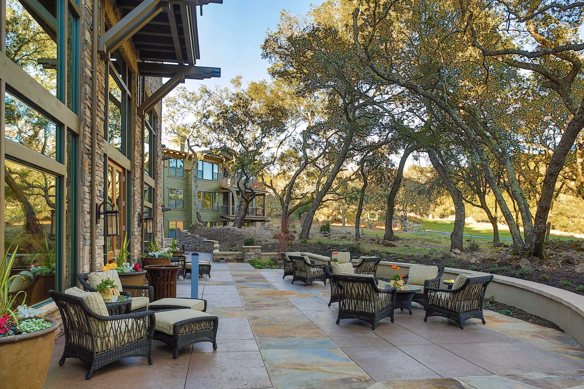 Outdoor patio area at Fountaingrove Lodge with cushioned wicker chairs and tables arranged on a tiled floor. The patio is adjacent to a building with large windows and stone walls. Surrounding the patio are trees and natural landscaping under a clear sky.