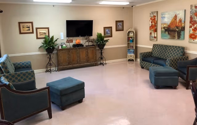 A bright seating lounge with upholstered chairs and ottomans, a TV over a wooden cabinet, potted plants and wall art.