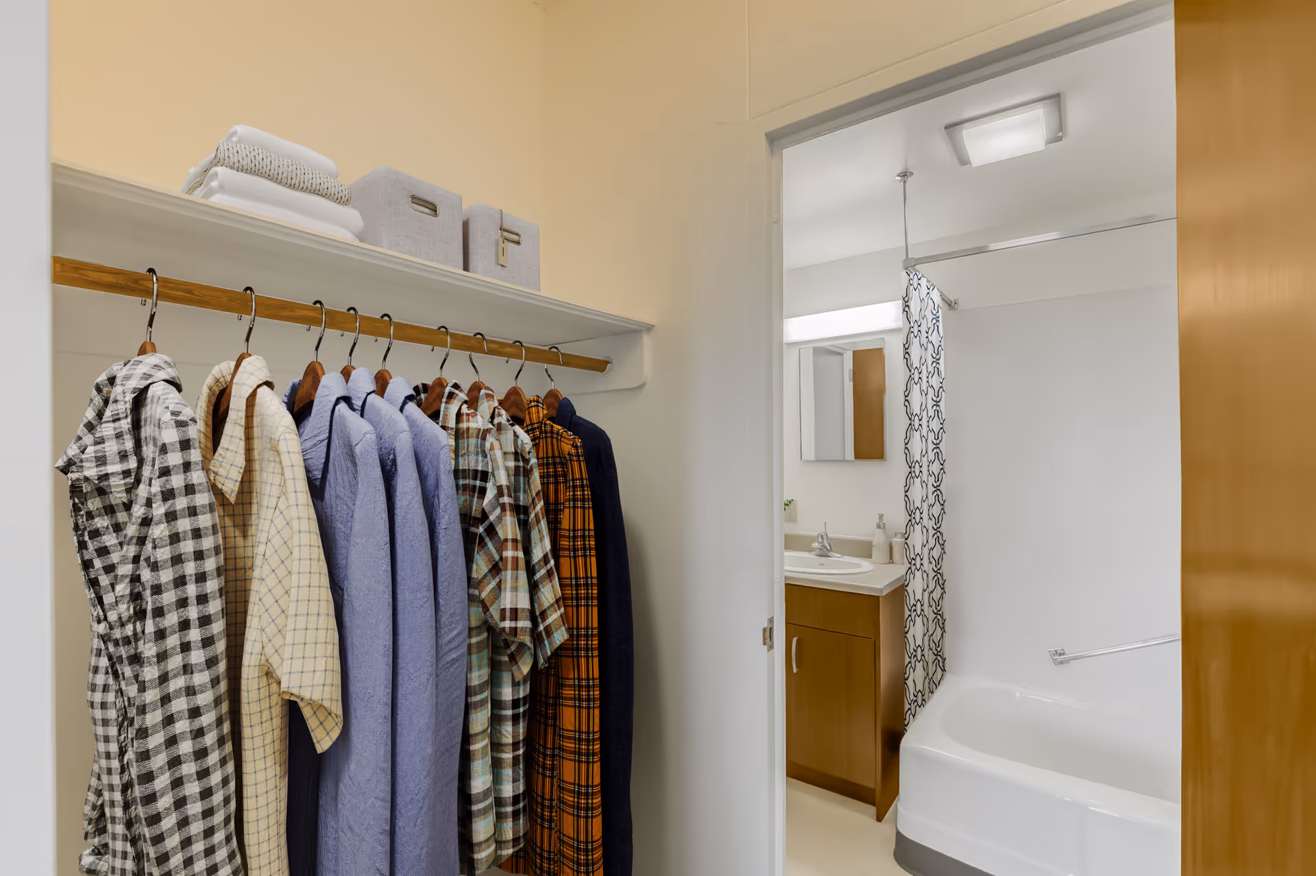 View of a closet with several shirts hanging on a wooden rod and folded towels and storage bins on the shelf above. The closet opens into a bathroom with a bathtub, shower curtain, sink, and mirror.