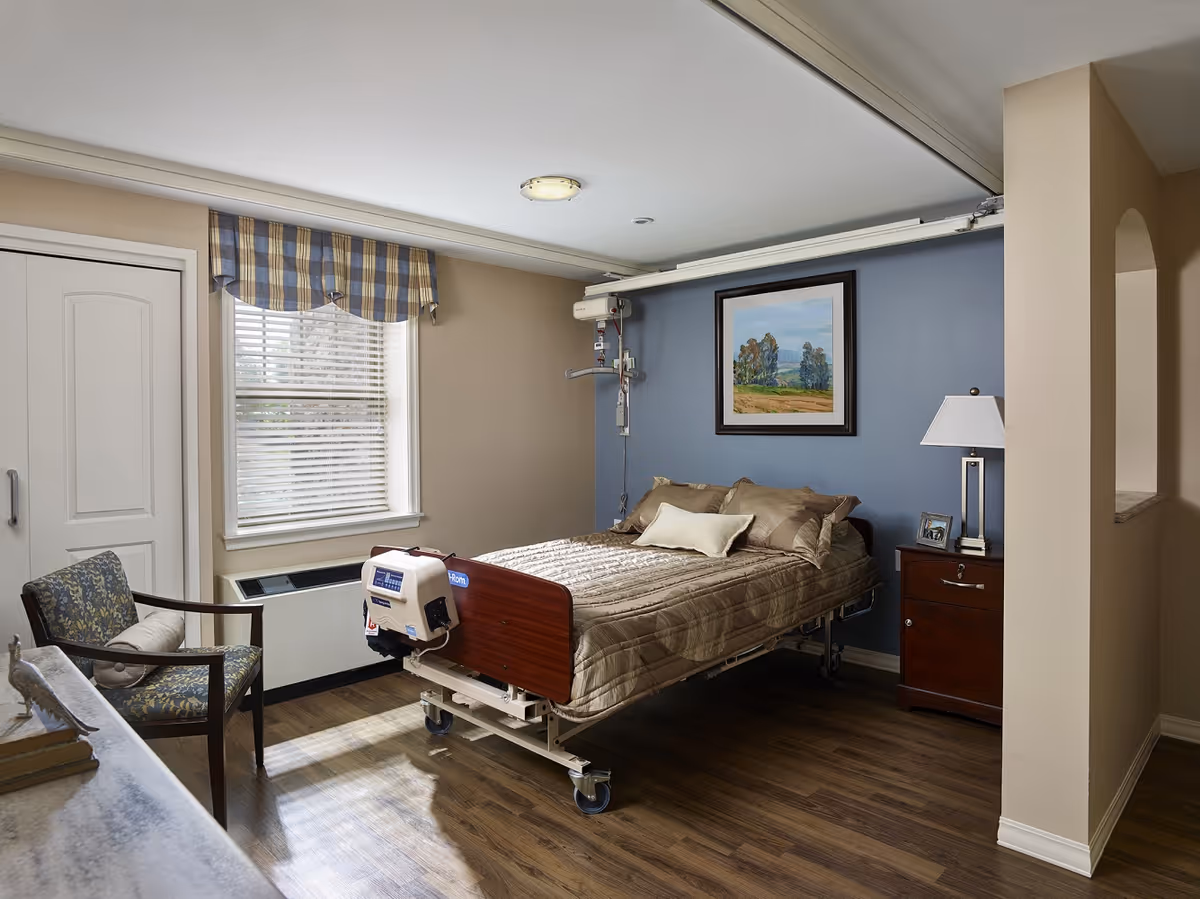 A well-lit bedroom in a senior living facility featuring a hospital-style bed with beige bedding against a blue accent wall. There is a framed landscape painting above the bed, a wooden nightstand with a lamp and a small photo frame, a window with blinds and a plaid valance, and a floral-patterned chair with a cushion. The floor is wood, and the room has neutral beige walls with a small archway visible on the right.