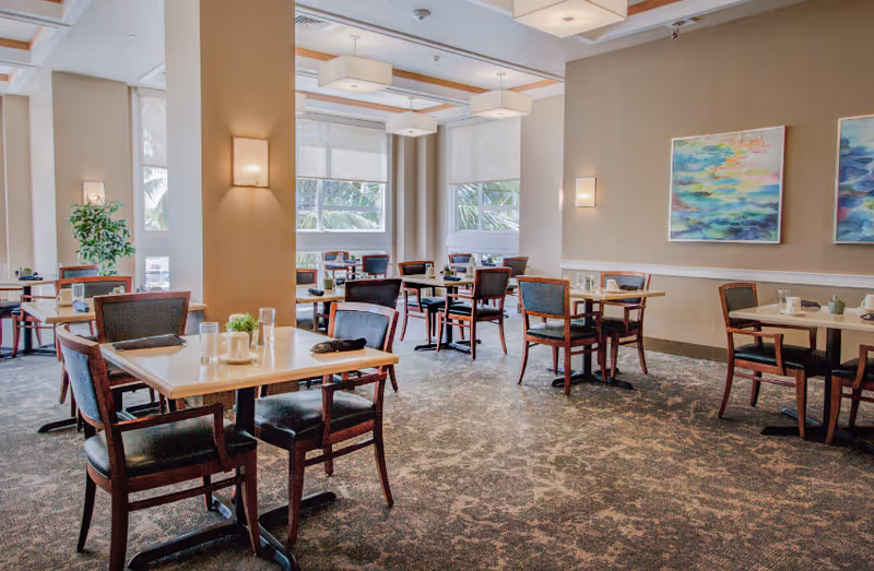 A bright dining room with several wooden tables and chairs arranged neatly. The room has large windows with white blinds, beige walls adorned with colorful abstract paintings, and modern ceiling lights. There are small plants and table settings on each table.