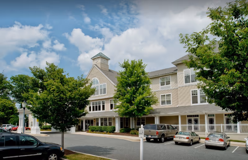 Two story senior living facility with beige siding, a parking lot, and green trees.