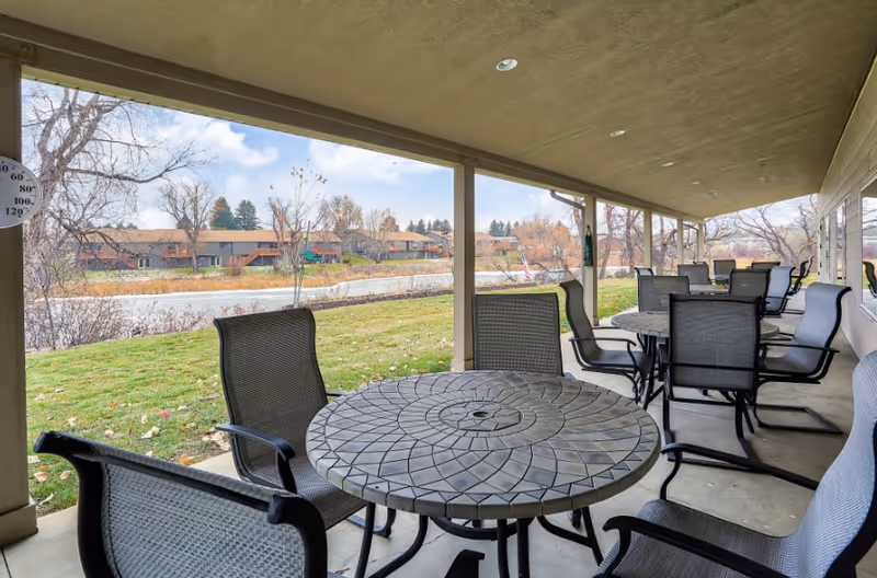 Covered outdoor patio area with multiple round mosaic tile tables and black mesh chairs. The patio overlooks a grassy area and a body of water with trees and residential buildings in the background under a partly cloudy sky.